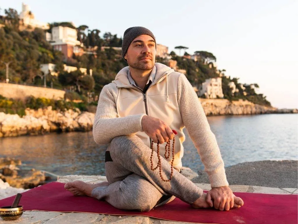 A man meditating, sitting on a red mat by the sea, wearing a beige hoodie, grey jeans and a grey cap, with a prayer bracelet in his hand, at sunset with a view of the coast and houses in the background.